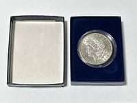 Photo showing the silver Morgan dollar coin housed in a protective plastic capsule and a blue presentation box next to a black outer cover.