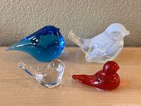 Four decorative glass birds displayed side by side on a wooden surface against a wall.