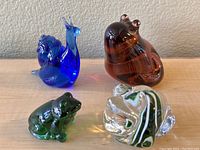 Four glass paperweights shaped like a blue snail, brown snail, green frog, and a clear swirled design on a light wood surface.