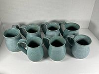 Photo showing full grouping of eight stoneware mugs with varying green-blue finishes and handles, arranged on white surface against white background.