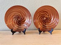 Two Murano glass plates displayed upright on black stands showing the red/brown swirl pattern with coppery sparkle.