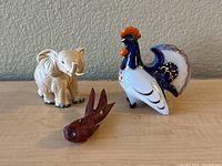 Three animal figurines on a neutral surface: a white elephant, a colorful rooster, and a small brown fish.
