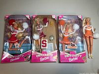 Four University Cheerleader Barbie dolls, three packaged and one loose, each in cheerleading outfits with pom-poms and university logos, displayed on a gray surface.