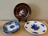 Five decorative plates including one maroon plate with gold rim and painted woman & child scene, four blue and white floral plates.