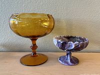 Two vintage glass candy dishes, amber and marbled purple-white, on pedestal bases displayed side by side.