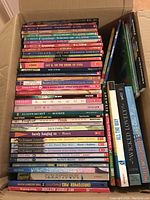 Stack of approximately 40 paperback books in a cardboard box, showing spines with titles and authors visible.