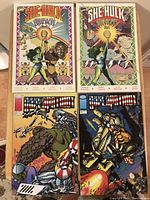 Four comic books laid flat on a table: two She-Hulk comics side by side on top, two Bloodstrike comics below them.