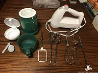 White electric hand mixer with five metal attachments (two beaters, one whisk, one dough hook, one flat beater) and green thermos with detachable cup and folding spoon.