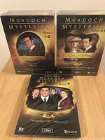 Two boxed DVD collections for Murdoch Mysteries Seasons 1-4 and 5-8 standing, with Season 9 collection laying flat on a wooden surface.