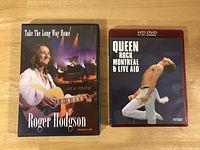 Two music DVDs side by side on a wooden surface. Roger Hodgson's Take The Long Way Home on the left with a purple stage background; Queen Rock Montreal & Live Aid HD DVD on the right featuring Freddie Mercury performing.