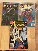 Three comic books laid flat on a wooden surface. Front covers visible showing Superman comics: Superman: Man of Steel #7, The Adventures of Superman #501, and Action Comics #667.