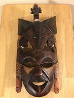 Front view of a dark wooden mask with detailed carvings across the face and a sitting figure on the forehead.