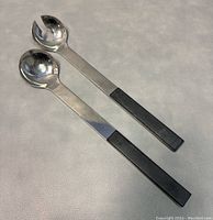 Pair of metal and black handled modernist Scandinavian salad tongs laying flat on light surface.