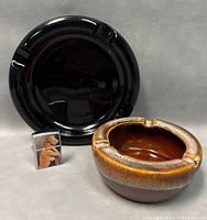 Photo of black glass ashtray, brown ceramic ashtray, and untested Supreme body tobacco lighter with risqué pinup girl image.