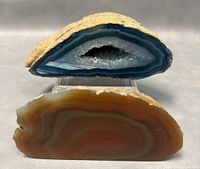 Close-up of the blue Inner Mongolia Gobi Eye agate geode showing the banding, crystal cavity, and rough exterior along with the polished reddish-brown agate slice in front.