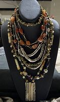 Multi-strand necklace displayed on a black mannequin featuring irregular stone beads in varied earth tones, silver-tone metal beads, and a large rectangular pendant.