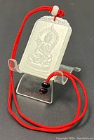 Front view of the white jade pendant showing seated deity carving and surrounding flame motif, displayed on clear stand with red cord and black/red beads attached.