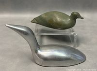 Photo showing two figurines: a green Inuit soapstone loon carving on a clear acrylic stand, and a silver aluminum Hoselton loon figurine in front.