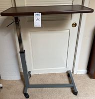 Full view of adjustable overbed table showing dark wood-effect tabletop and gray metal base with wheels.