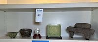 Full display of all objects on shelf from left to right: small green bowl, bronze bowl, painted egg on stand, green wax cube candle, stone grinder set
