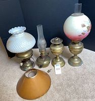 Four brass lamps including two oil lamps with glass chimneys, one electric lamp and one small brass lamp base. Included are two glass lamp shades (white hobnail with ruffled top, floral painted globe) and one separate brown lamp shade.