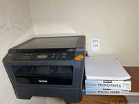 Black Brother HL-2280DW wireless laser printer with two unopened boxes of Kirkland Professional Glossy Inkjet Photo Paper stacked beside it.