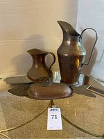 Three metal items displayed together: a tall hammered brass pitcher with a high curved handle, a shorter rounded copper pitcher with a single handle, and a fish-shaped decorative copper bowl with a lid and fin handle.
