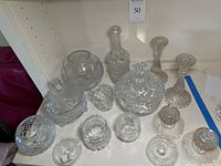 Wide view showing multiple cut glass and crystal items including spherical vases, footed bowls with lids, various small jars, decanter without top, and candlesticks.
