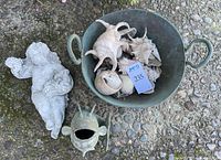 Concrete cherub figure with grape and leaf decoration, metal bowl with seashells, metal fish sculpture on pebble surface