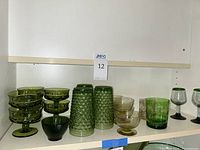 Shelf with six green stemmed parfait cups, five yellow/green sherbet cups, three heavy pressed bubble textured green drinking glasses, one thick textured glass bowl, and two short brown-tinted glasses.