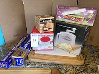 Photo showing all the kitchen gadgets including boxed microwave rice cooker, Donvier ice cream maker, terracotta garlic oven, Vidalia vegetable chopper, cutting boards, and unopened foil and paper rolls