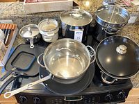 Cookware items displayed on stove including white Crêpe Master set, small square griddle, stainless steel sauce pan, and black roasting pan