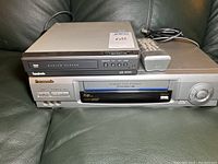 Panasonic VCR below Symphonic DVD/CD player with remote on green leather couch