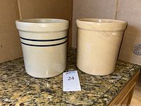 Two ceramic crocks standing side by side on granite countertop with a lot number card in front.