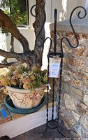 Photo showing the wrought iron plant stand with its twisted legs and curled hooks standing next to a faux stone pot filled with artificial succulents, placed outside against a stone wall.