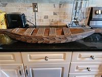 Front view of hand carved wooden canoe placed on kitchen counter with visible leaf pattern carvings and multiple raised wooden slat seats bound with natural twine.