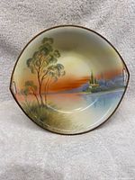 Front view of bowl showing hand painted landscape with trees, grass, water, and buildings at sunset in warm colors.