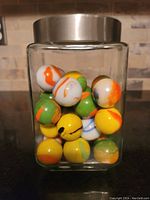 Clear glass jar filled with large multicolored marbles, showing orange, yellow, green, white, and blue colors with various swirled patterns.