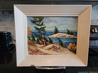 Frontal photo of original oil painting showing detailed landscape scene with rocky shore, bent pine trees, and distant island in blue water. White frame with wear is visible.