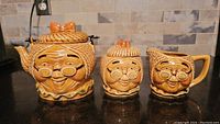 Full view of tea pot, sugar bowl, and creamer all designed with grandmother face motifs in a warm caramel colored ceramic.