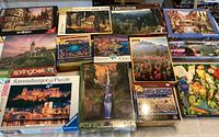 Full lot of 13 jigsaw puzzle boxes laid out on table, showing varied scenes of lighthouses, towns, landscapes, and art-inspired images.