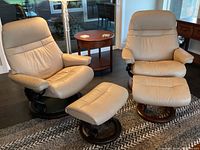 Both beige leather recliner chairs and ottomans displayed side by side on floor mat, wood finishes visible.