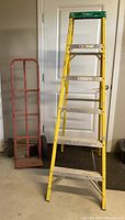 Yellow Werner 6-foot ladder next to red heavy duty hand truck dolly, positioned upright indoors near a white door.