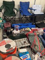 Two foldable camping chairs, cooler, portable stove, bucket toilets, sleeping pads, camping tools and supplies arranged on floor
