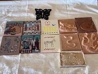 Full view of all nine southwestern themed ceramic tiles and one cast iron butterfly trivet on white cloth.
