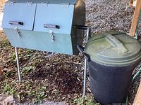 Left-front view of green metal compost tumbler standing on four legs next to black plastic trash can