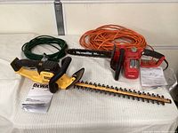 DeWalt cordless hedge trimmer laid flat with Homelite electric chainsaw and two extension cords visible behind. User manuals included.