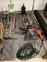 Photo of garden tools arranged on a tarp showing various rakes, shovels, pruning tools, plastic shears, a sprayer, and gardening supplies including bags and jugs.