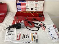Electric Milwaukee Sawzall in red with attached cord, assorted blades laid in front, red metal carrying case behind all items