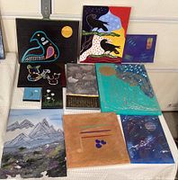 Overview of 11 paintings showing variety of subjects and sizes arranged on table.